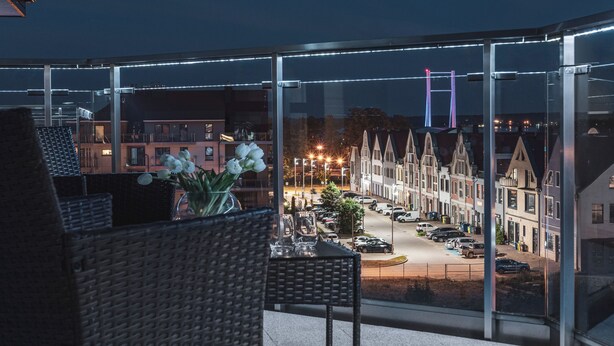 A balcony with wicker furniture, a vase with flowers, and a view of a street with parked cars and buildings. - Apartamenty Sun & Snow Róża Wiatrów (Gizycko)