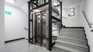A modern elevator with a glass door, a staircase with a metal railing, and a large number '3' on the wall. - Apartamenty Sun & Snow Róża Wiatrów (Gizycko)