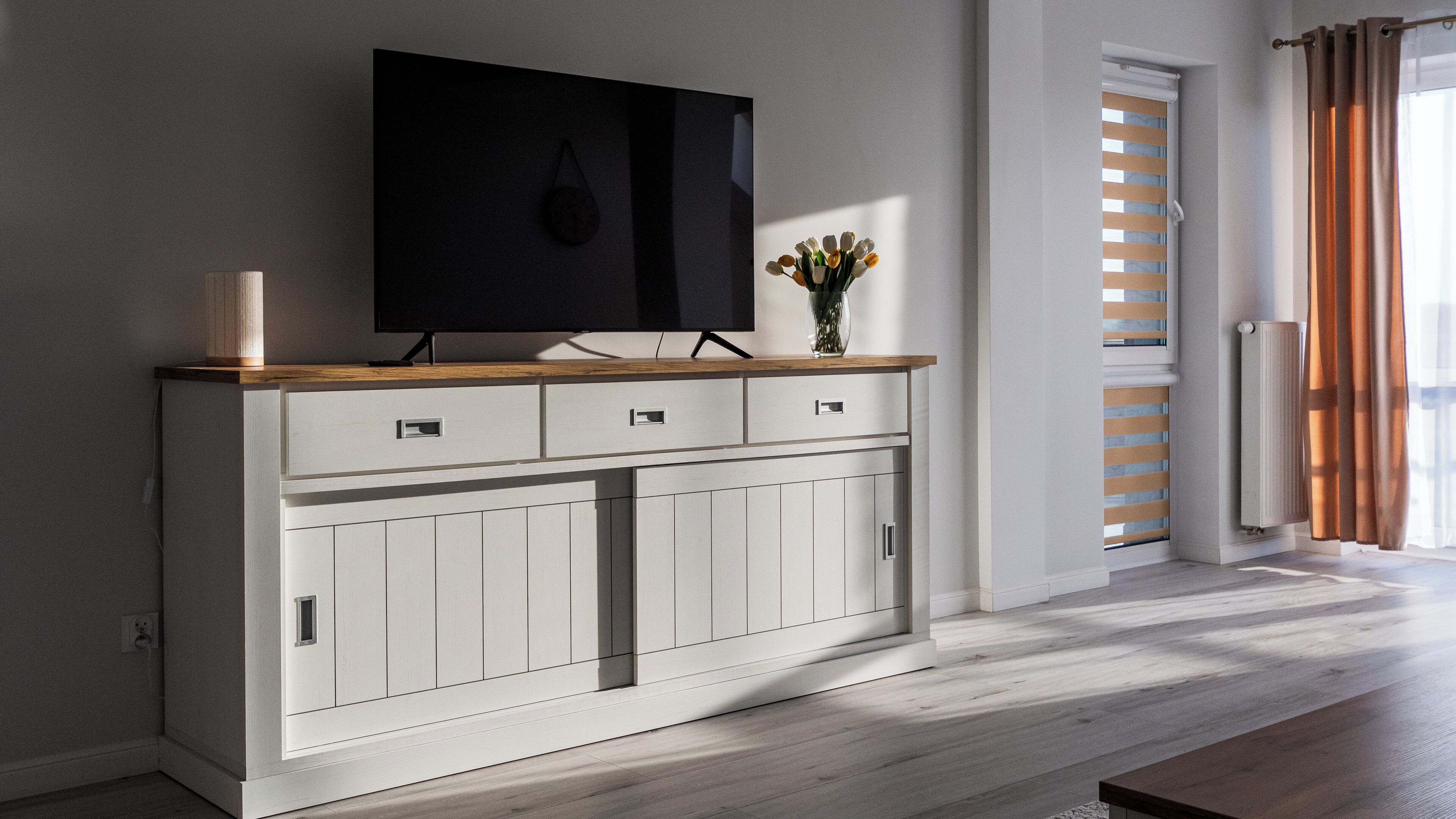 A modern living room with a flat-screen TV on a wooden console, a vase with flowers, and a view of the outdoors through a window with blinds.