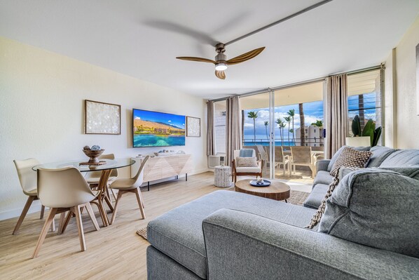 A modern living room with a grey sofa, a dining area with a glass table, and a balcony with a view of the ocean. - Beautiful Oceanfront Condo, Huge Views near Harbor, AC! (Wailuku)