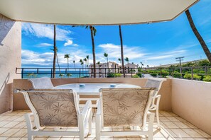 A balcony with a round table and chairs, overlooking a beach with palm trees and a clear sky. - Beautiful Oceanfront Condo, Huge Views near Harbor, AC! (Wailuku)
