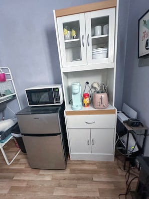 A compact kitchenette with a refrigerator, microwave, kettle, toaster, and a cabinet with dishes.