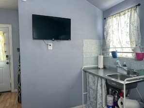 A compact kitchen with a flat-screen TV mounted on the wall, a sink with a stainless steel countertop, and a window with floral curtains. - TH Monthly + Utilities + Laundry + Comfy Stay! (Grant)