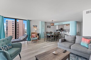 Un salon moderne avec un canapé, un fauteuil et une table basse. On y trouve également un coin repas avec une table et des chaises. La pièce est dotée d’une grande fenêtre avec des rideaux.