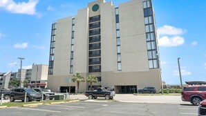 A multi-story building with a central vertical feature, parking lot, and adjacent smaller structures. - Bright & Spacious Direct Oceanfront 3BR Condo in North Myrtle Beach! (North Myrtle Beach)