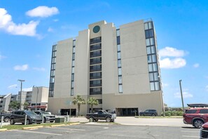 A multi-story building with a central vertical feature, parking lot, and adjacent smaller structures.