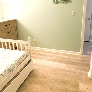 A room with a wooden floor, a white crib, a dresser with drawers, and a wall socket.