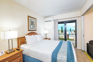 A hotel room with a bed, a nightstand, a lamp, a framed picture, and a view of a pool and palm trees through the door. - Emerald Seas Deerfield Beach Club (Deerfield Beach)