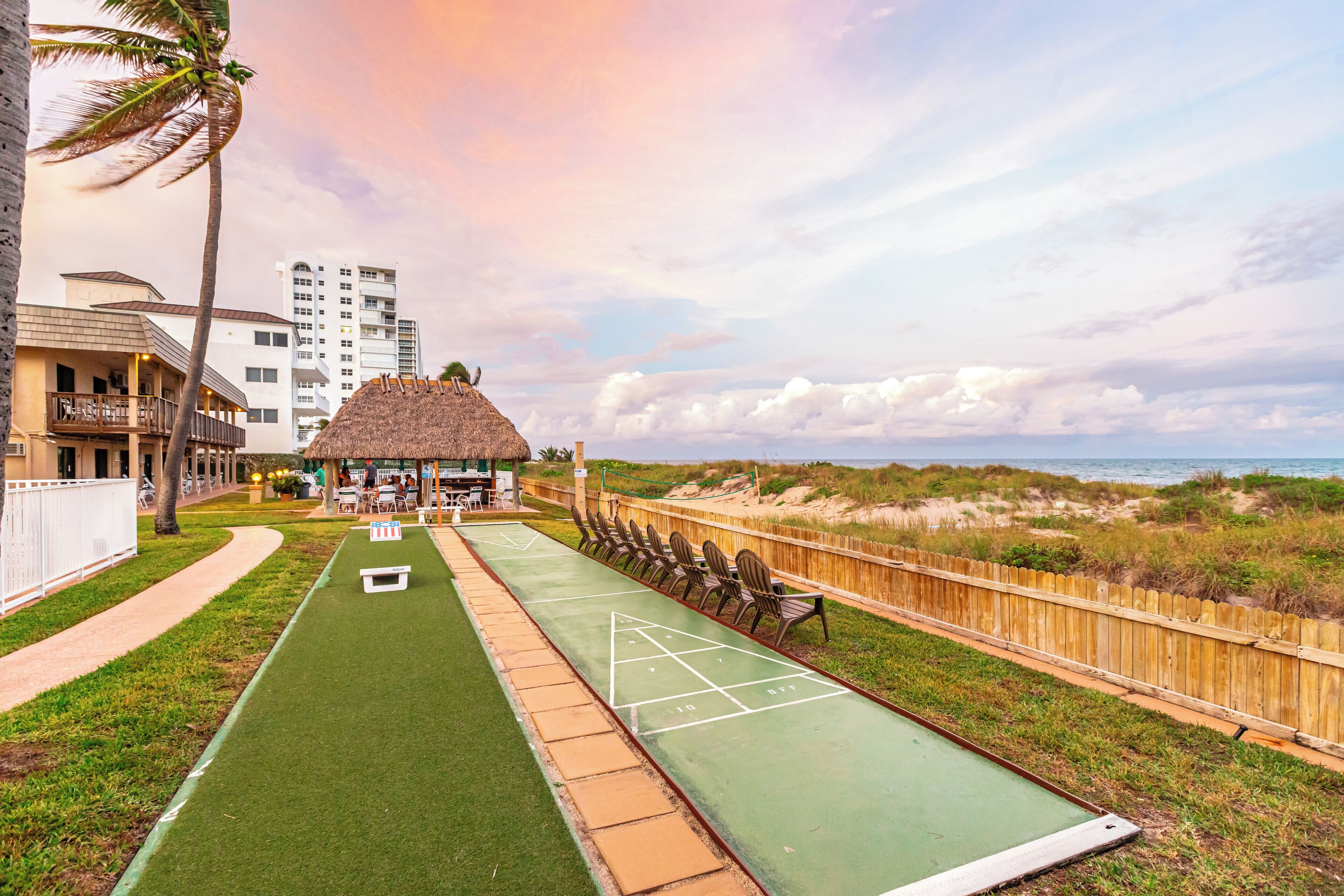 A shuffleboard court with a wooden fence, a thatched-roof structure, and a view of the beach and buildings in the background.