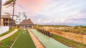 A shuffleboard court with a wooden fence, a thatched-roof structure, and a view of the beach and buildings in the background.