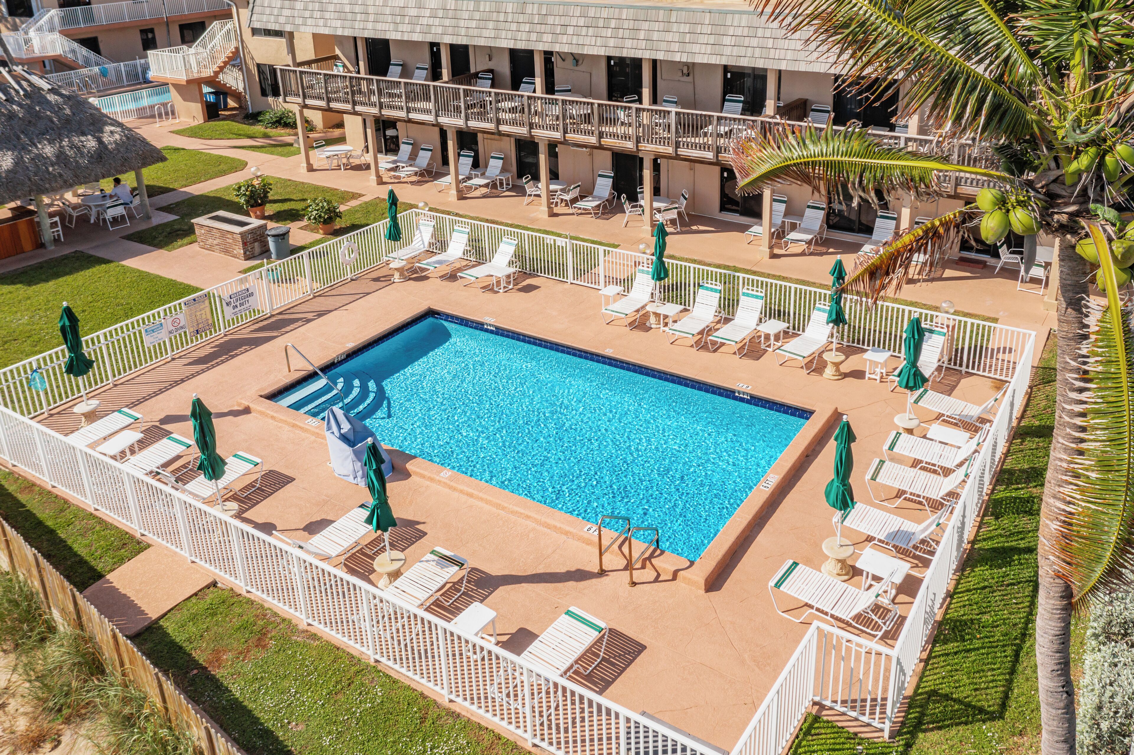 Zona de piscina con sillas de descanso y sombrillas, rodeada por una valla blanca.