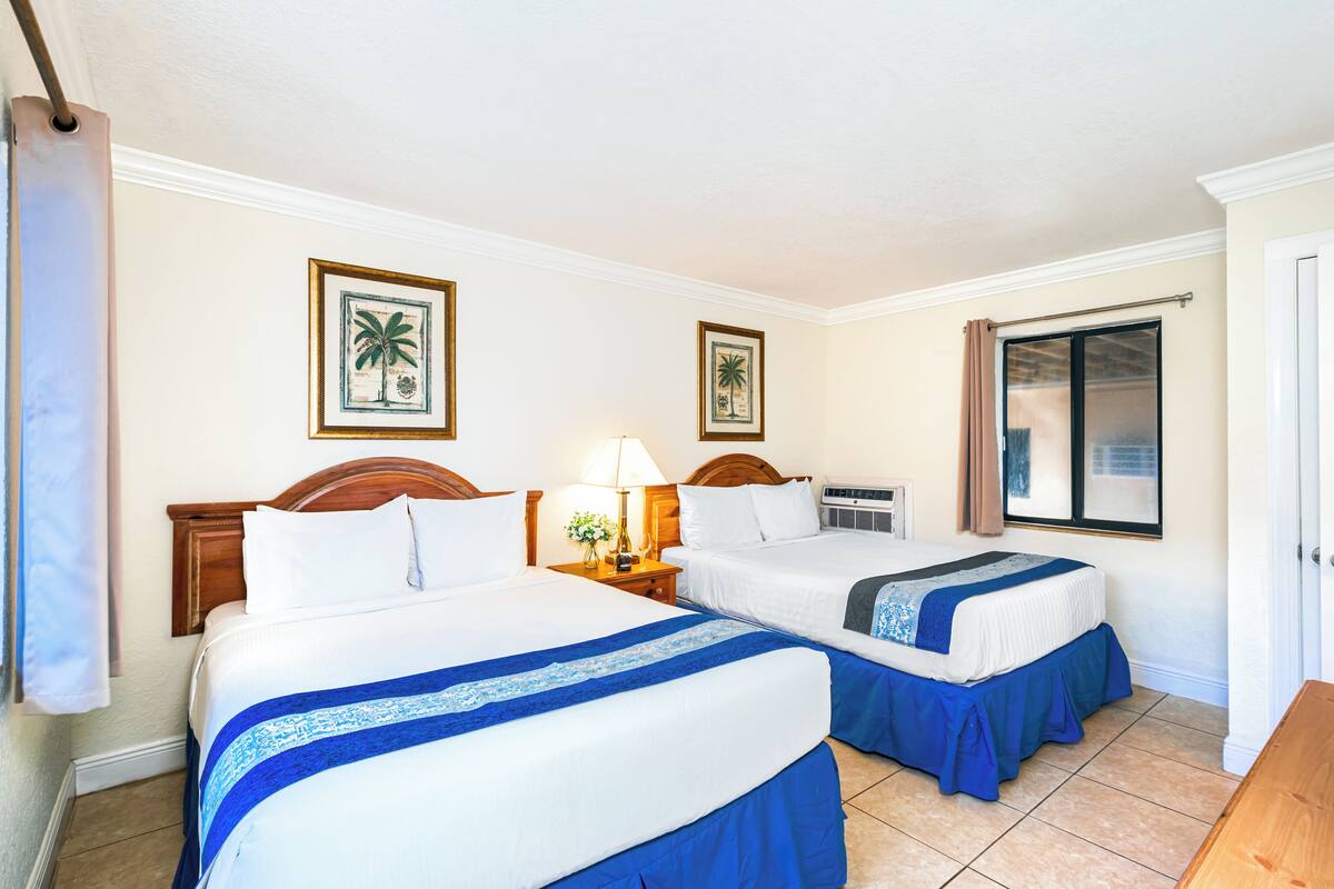 Ein Hotelzimmer mit zwei Betten, einem Nachttisch mit Lampe, gerahmten Bildern an der Wand und einem Fenster mit Vorhängen.