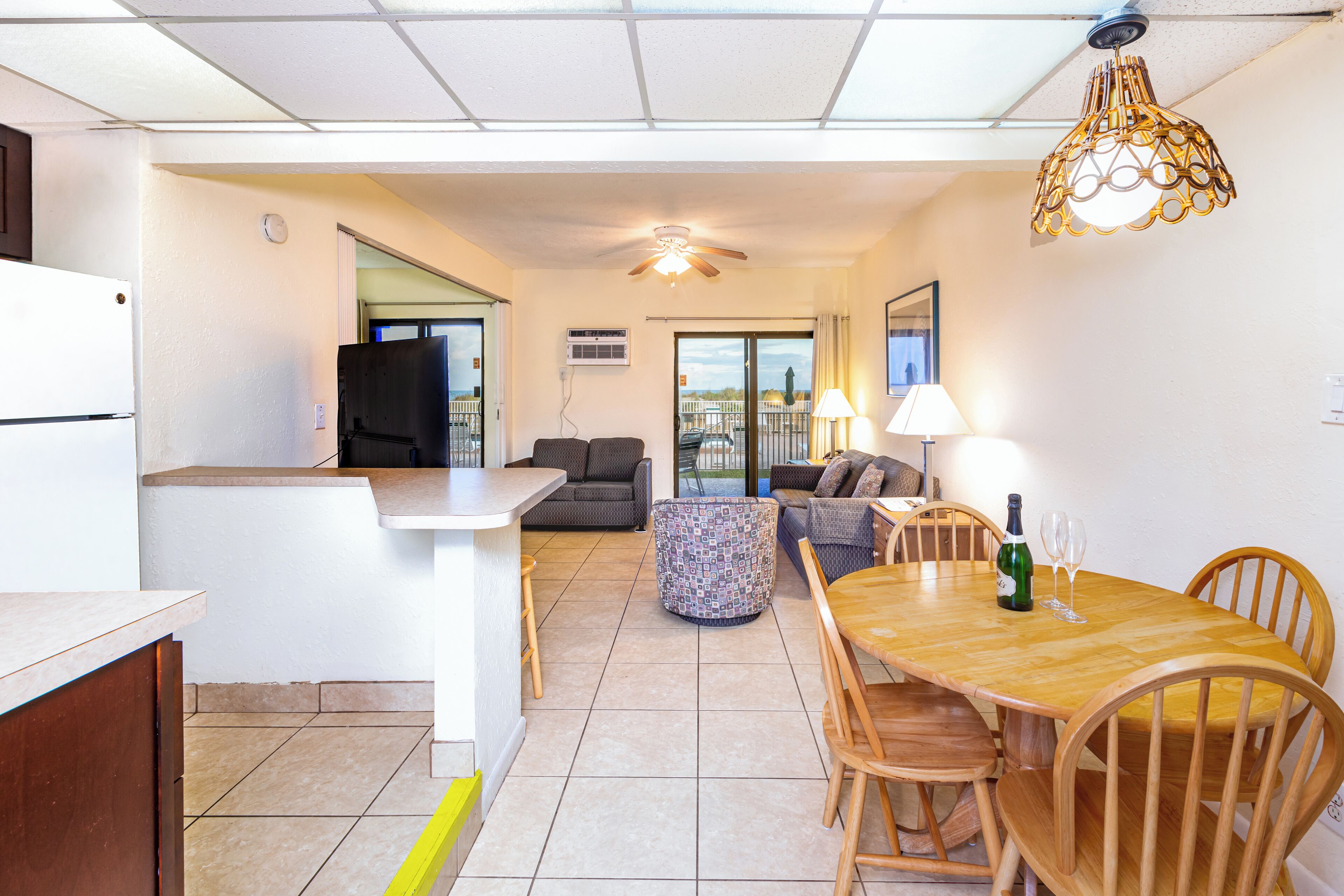 A modern kitchen with a white refrigerator, a dining area with a wooden table and chairs, a patterned ottoman, and a ceiling fan.