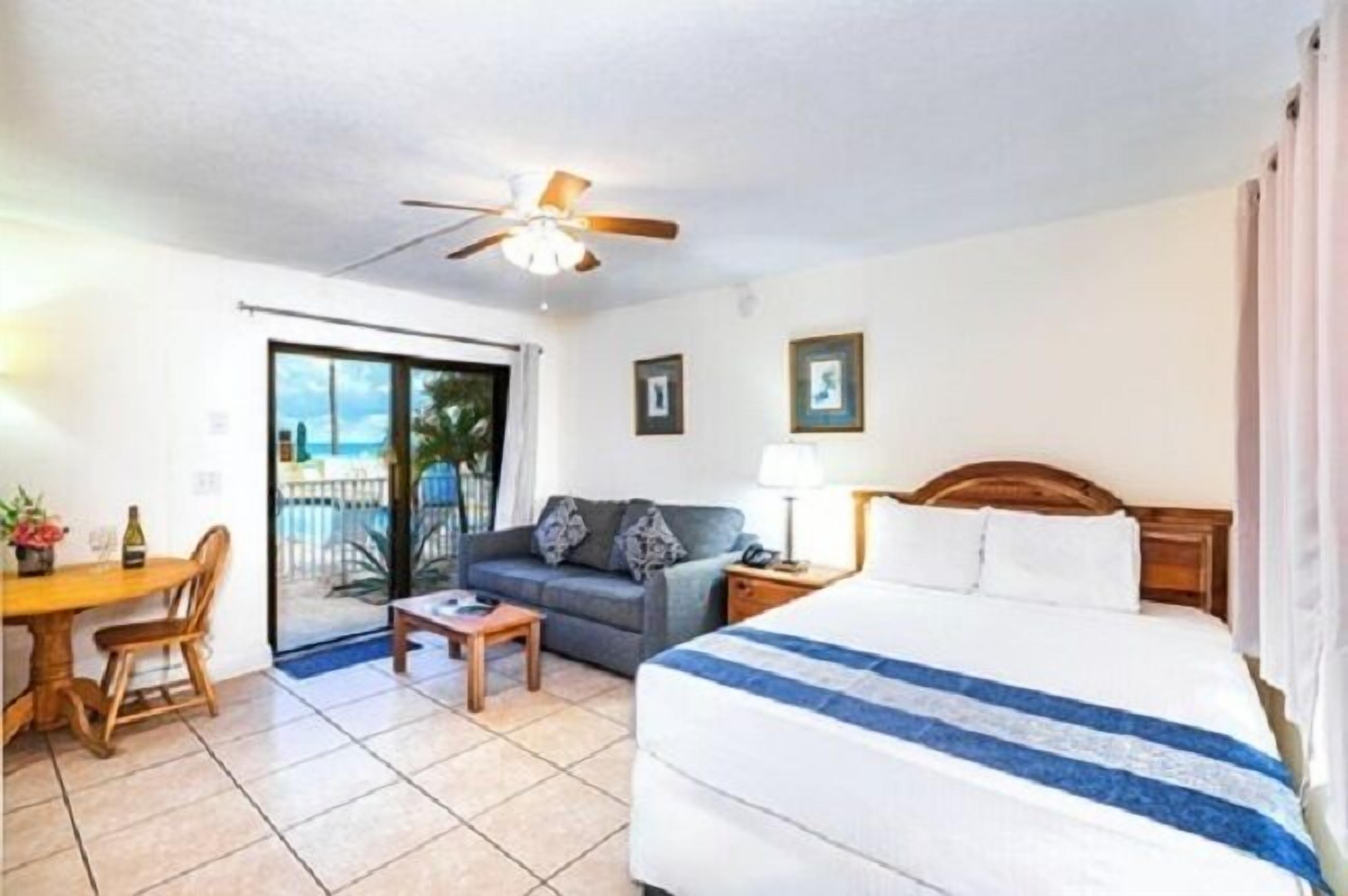 A hotel room with a bed, a sofa, a table, a chair, a ceiling fan, and a view through a sliding door to the outside.