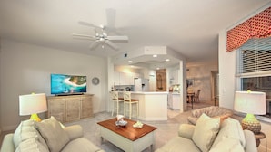 A modern living room with a sofa, coffee table, and TV. A kitchen and dining area are visible in the background. - Whisper Trace at Fiddlers Creek - 2 Bedroom Coach Home. (Naples)
