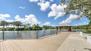 A wooden deck with a gazebo overlooking a calm body of water, surrounded by lush greenery and a clear blue sky. - Whisper Trace at Fiddlers Creek - 2 Bedroom Coach Home. (Naples)