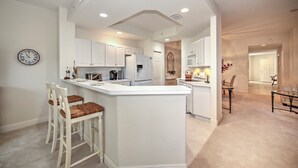 A modern kitchen with white cabinetry, a central island, and a dining area with a glass table. - Whisper Trace at Fiddlers Creek - 2 Bedroom Coach Home. (Naples)