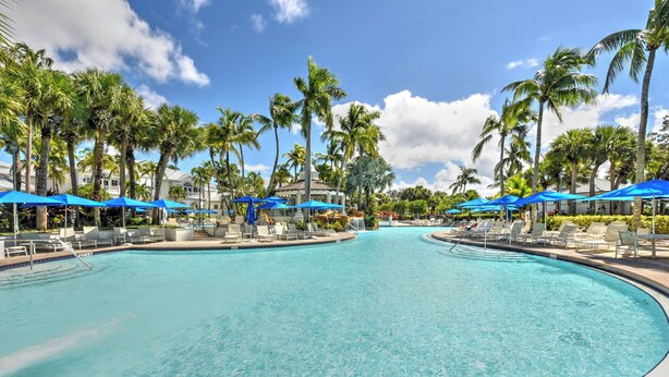 A large, clear swimming pool with blue umbrellas and lounge chairs, surrounded by palm trees and a clear sky. - Whisper Trace at Fiddlers Creek - 2 Bedroom Coach Home. (Naples)
