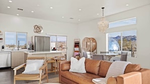 A spacious living area with a leather sofa, wicker chairs, and a view of a mountainous landscape through large windows. - Quail Wash Retreat: Star Dome, Sauna, Plunge, Spa (Joshua Tree)