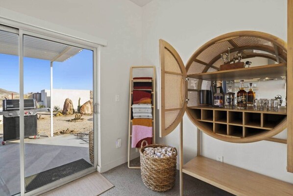 A room with a sliding glass door leading to an outdoor area, a woven basket, and a wooden shelf with bottles and glasses.