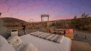 A room with a bed, a table, a chair, a telescope, and a view of the desert through a hexagonal window. - Quail Wash Retreat: Star Dome, Sauna, Plunge, Spa (Joshua Tree)