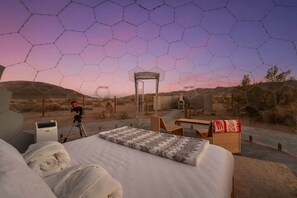 Ein Zimmer mit Bett, Tisch, Stuhl, einem Teleskop und Blick auf die Wüste durch ein Sechseckfenster.