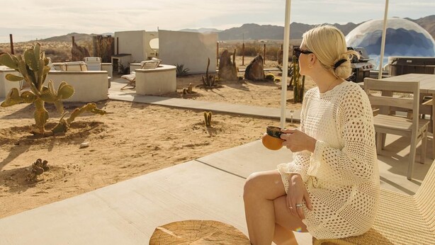 A person seated on a lounge chair in a desert setting with cacti, outdoor furniture, and a clear view of the landscape. - Quail Wash Retreat: Star Dome, Sauna, Plunge, Spa (Joshua Tree)