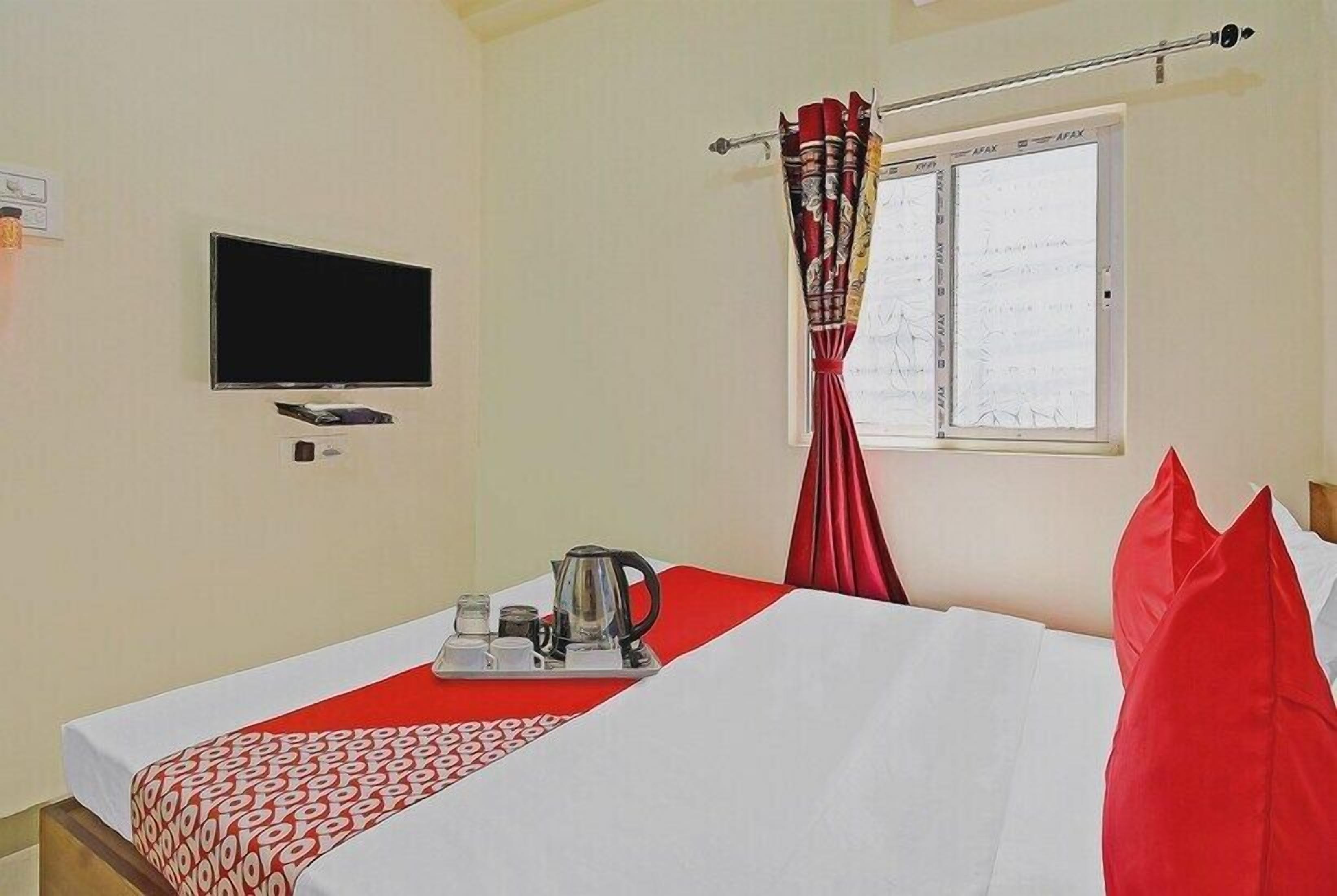 A hotel room with a bed, a TV mounted on the wall, a window with curtains, and a tray with a kettle and cups on the bed.