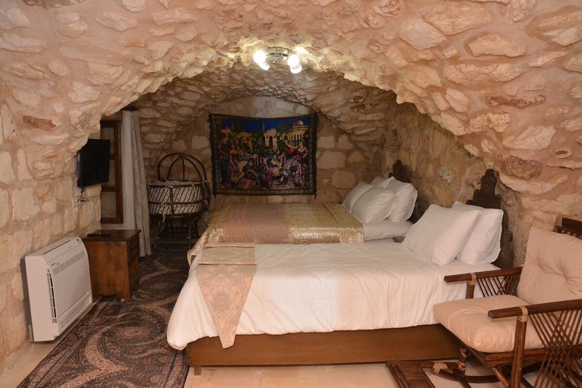 Una habitación acogedora con techo de piedra, una cama con sábanas blancas, una silla de madera y un tapiz en la pared.