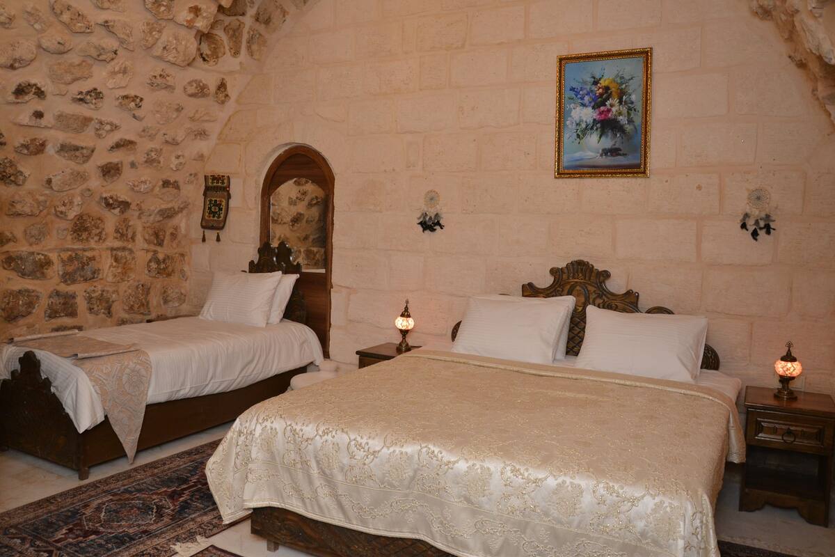 Una habitación con dos camas, paredes de piedra, una puerta de madera, un cuadro y una mesita de noche con una lámpara.