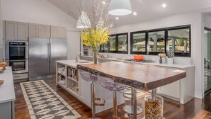 A modern kitchen with stainless steel appliances, a large island with bar stools, and a view through large windows. - Calistoga Wine Ranch Estate (Calistoga)