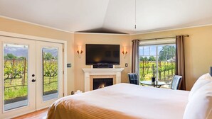 A hotel room with a bed, a fireplace, a TV, and a view of a vineyard through large windows.