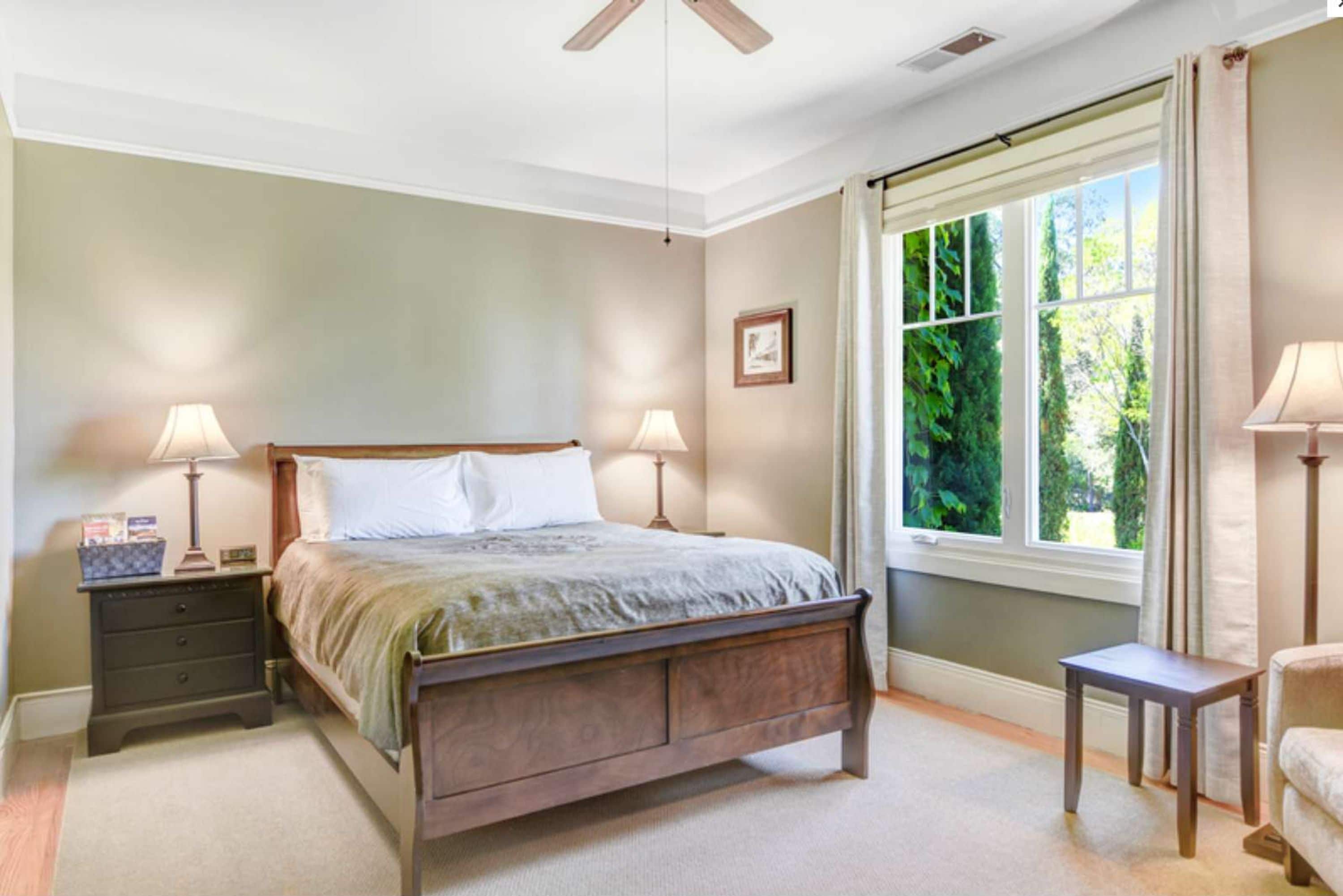 A bedroom with a wooden bed, two bedside tables with lamps, a window with a view of greenery, and a ceiling fan.