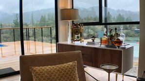 En moderne stue med et stort vindue der kigger ud på en terrasse og bjerge. Der er en sofa med et mønstret pude, et sofabord med en lampe og et skab med flasker og et ur.