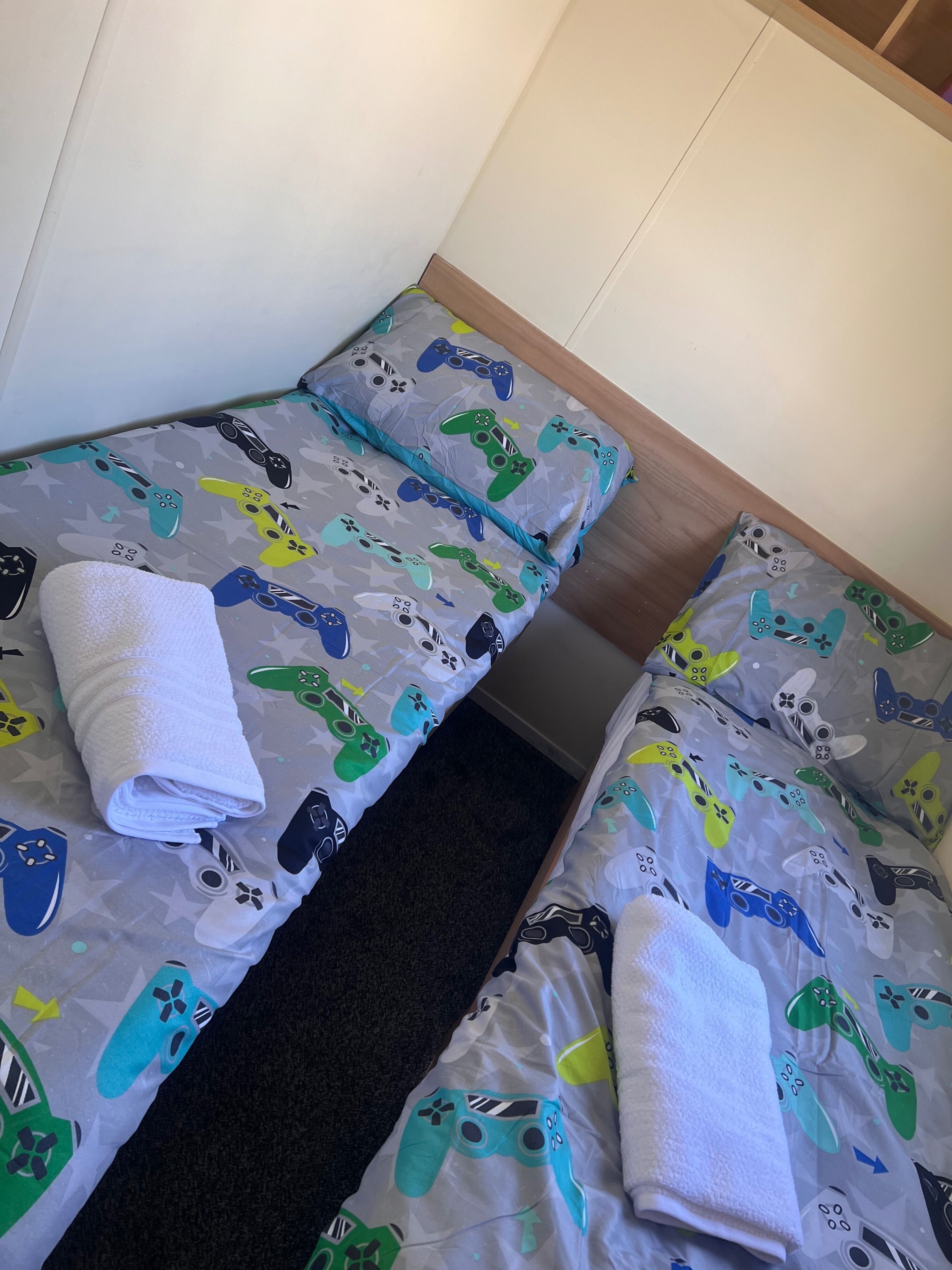 Two single beds with grey bedding featuring colorful game controller prints, a wooden headboard, and a dark carpet runner.