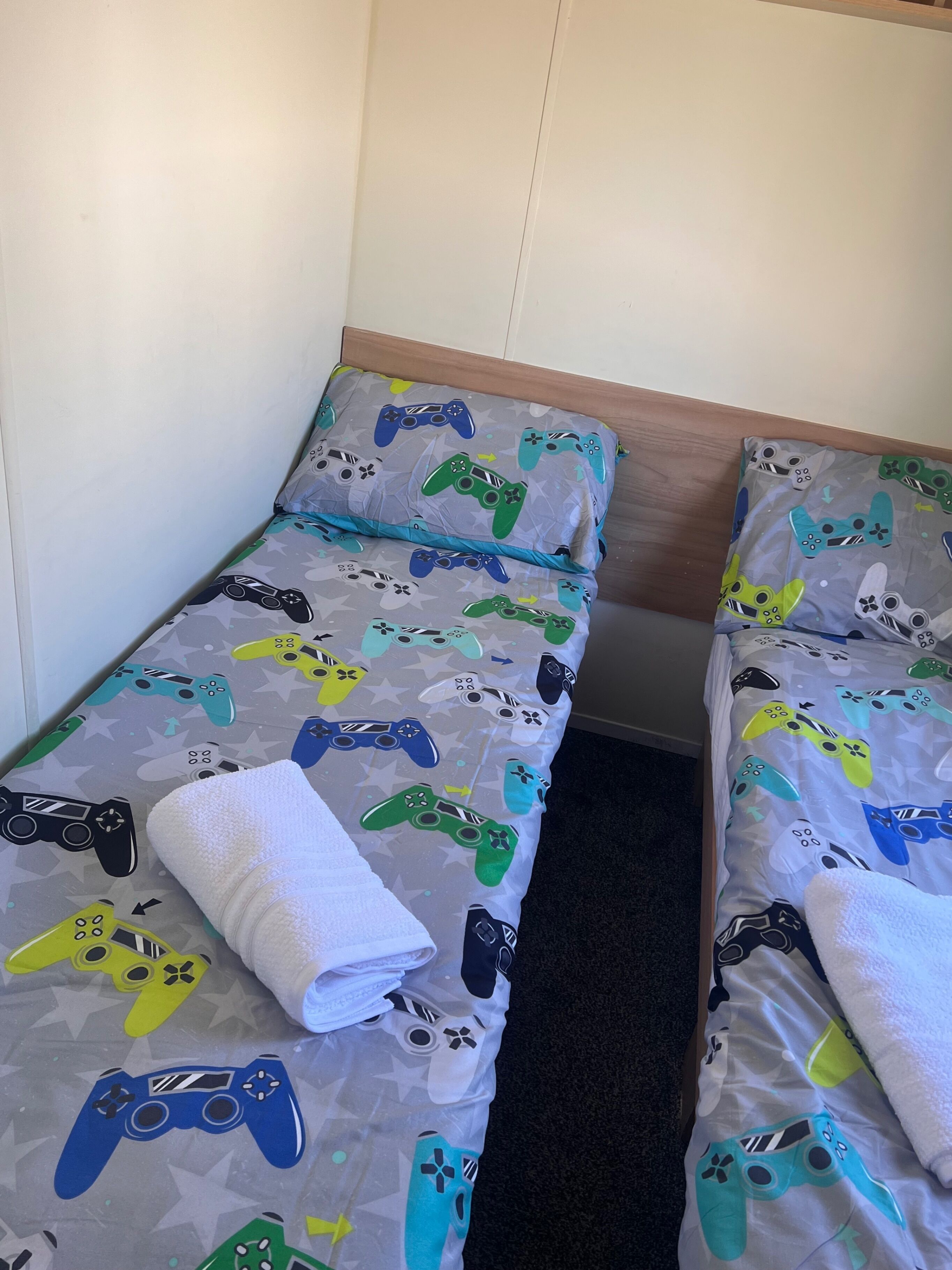 Two single beds with grey bedding featuring colorful game controller prints, white pillows, and a wooden headboard.