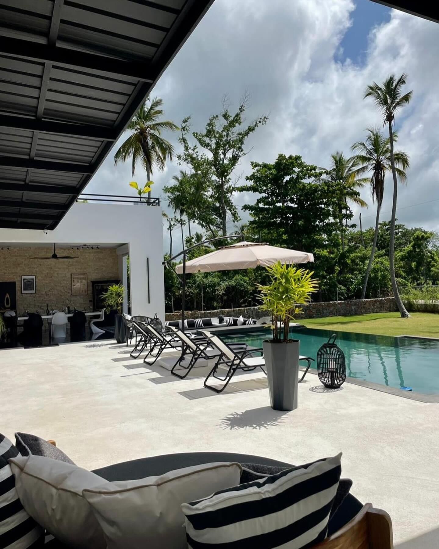 An outdoor pool area with lounge chairs, a covered seating area, and a view of palm trees and a building in the background.
