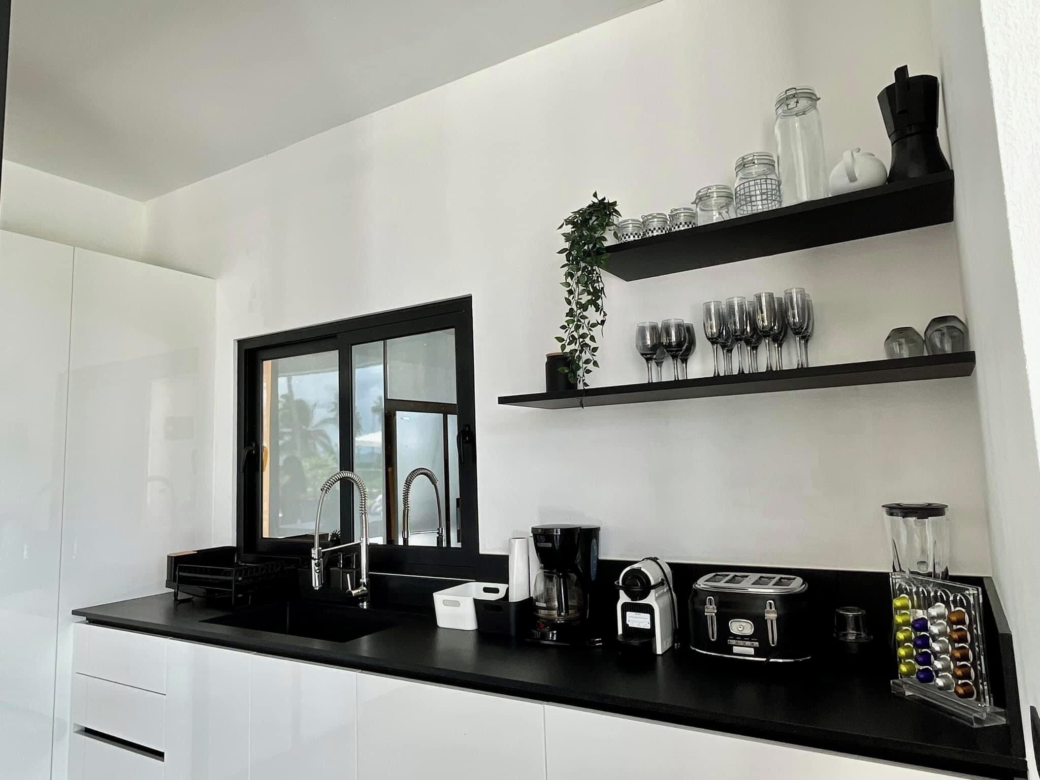 A modern kitchen with white cabinets, a black countertop, a window, and shelves with glassware and a coffee maker.