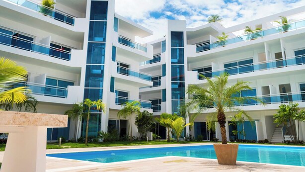 A modern multi-story hotel with a swimming pool, balconies, and palm trees. - Baya Azul Bayahibe - Penthouse 402 (San Rafael del Yuma)