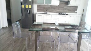 A modern kitchen with white cabinets, a stainless steel refrigerator, a glass dining table with four chairs, and a wooden backsplash. - Baya Azul Bayahibe - Penthouse 402 (San Rafael del Yuma)