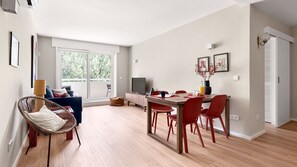 Ein modernes Wohnzimmer mit einer Couch, einem Esstisch mit Stühlen, einem Fernseher und einem großen Fenster mit Blick auf Bäume.