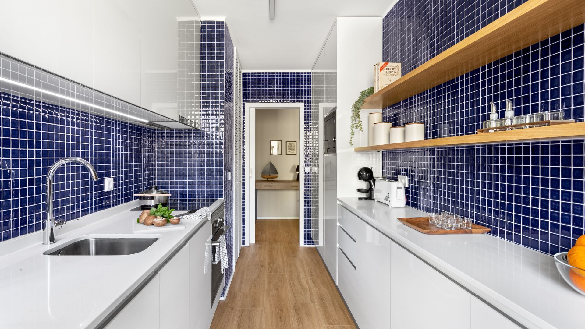 Une cuisine étroite aux murs recouverts de carreaux bleus, avec des plans de travail blancs et un plancher en bois.