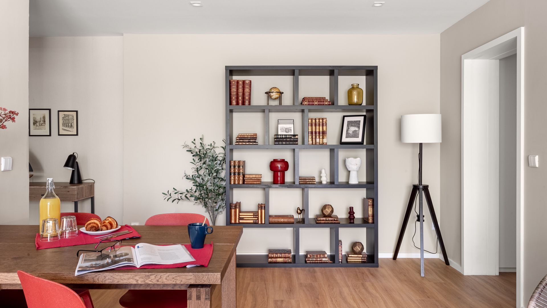 Ein modernes Wohnzimmer mit einem Holzesstisch, roten Stühlen, einem Bücherregal mit Büchern und Dekoration sowie einer Stehlampe und einem Blumentopf.