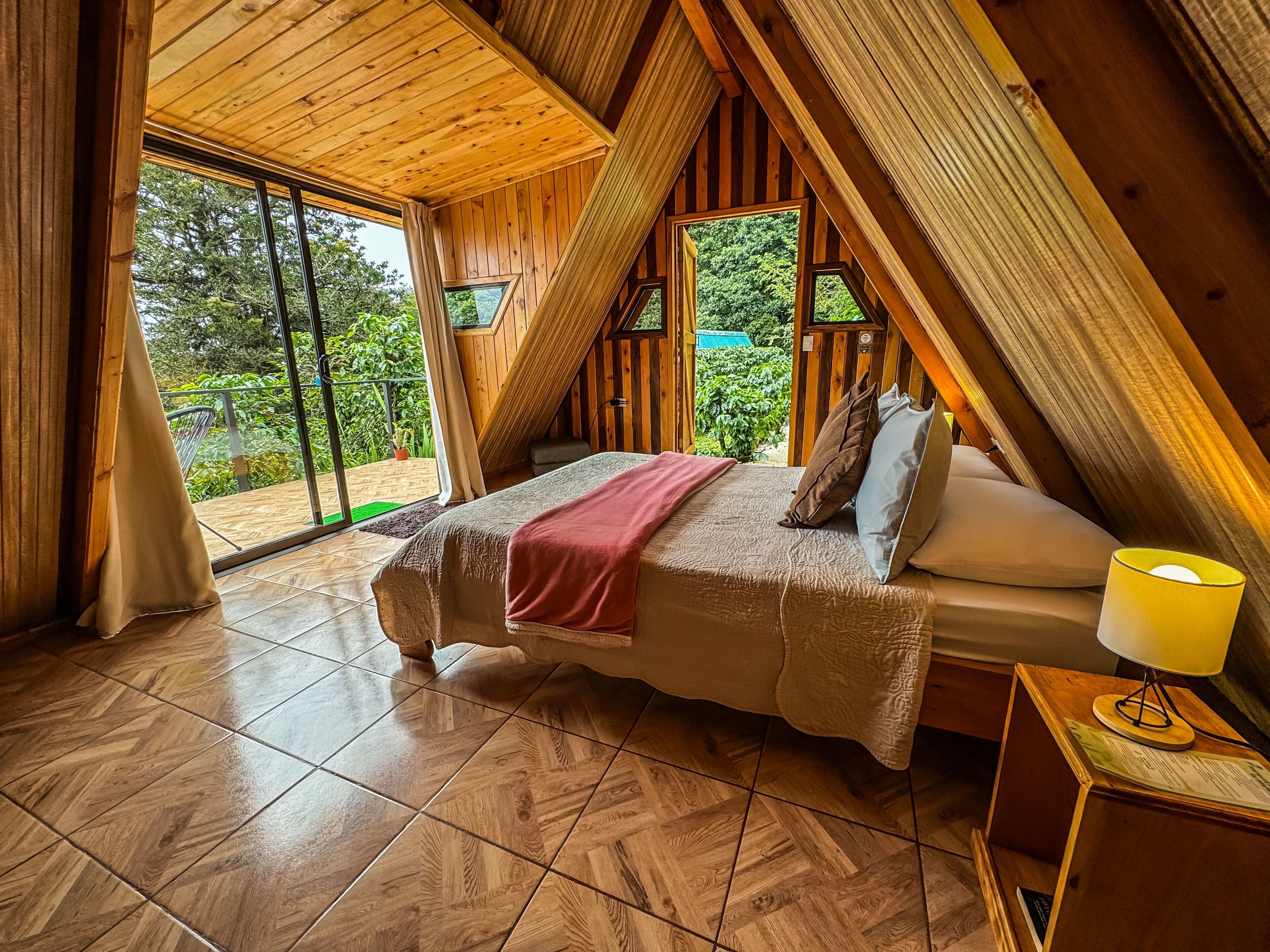 Quarto com teto inclinado de madeira, cama com almofadas, mesa de cabeceira com abajur e vista para o verde lá fora.