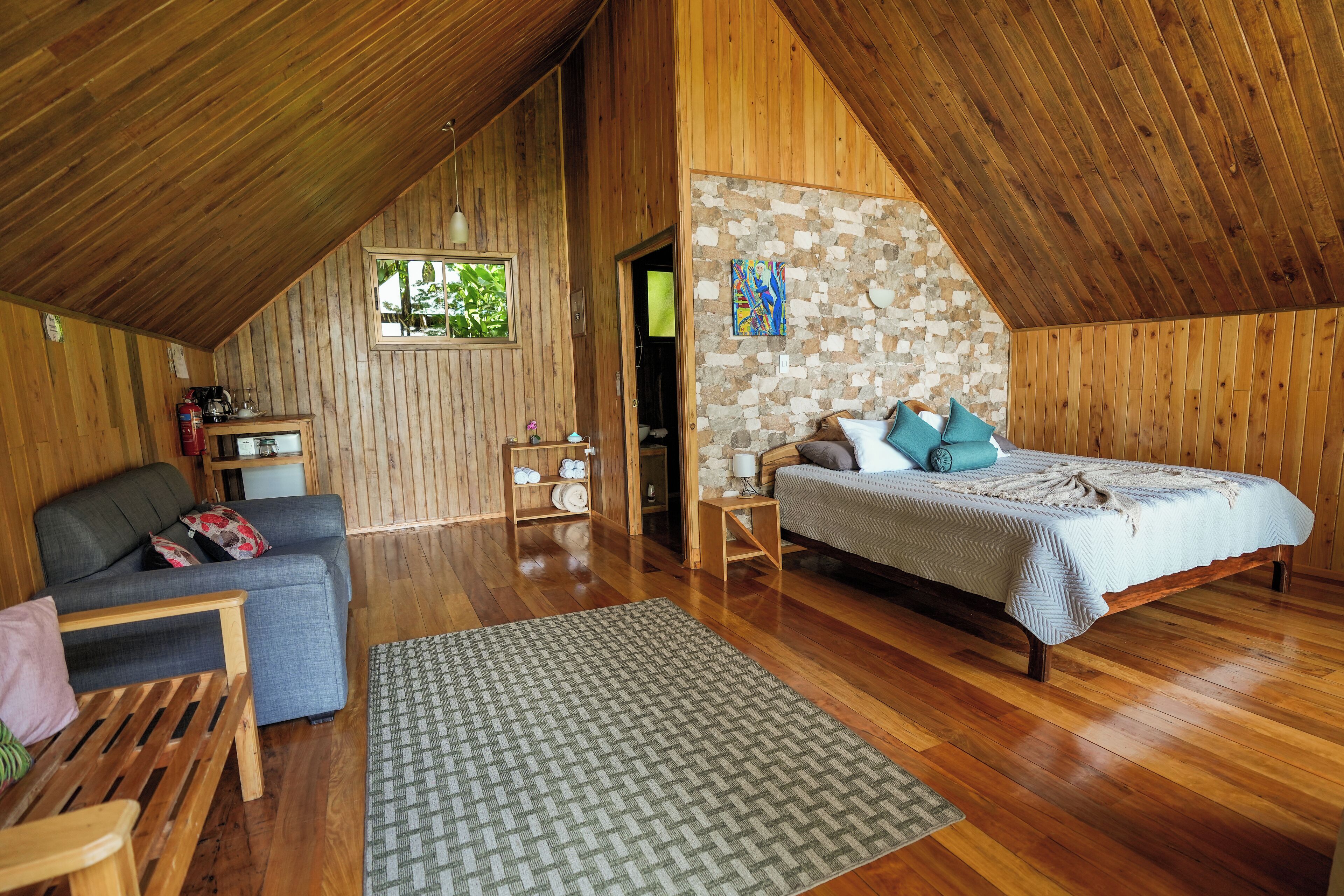 A cozy room with a wooden ceiling, a bed with pillows, a sofa, a small table, and a rug.