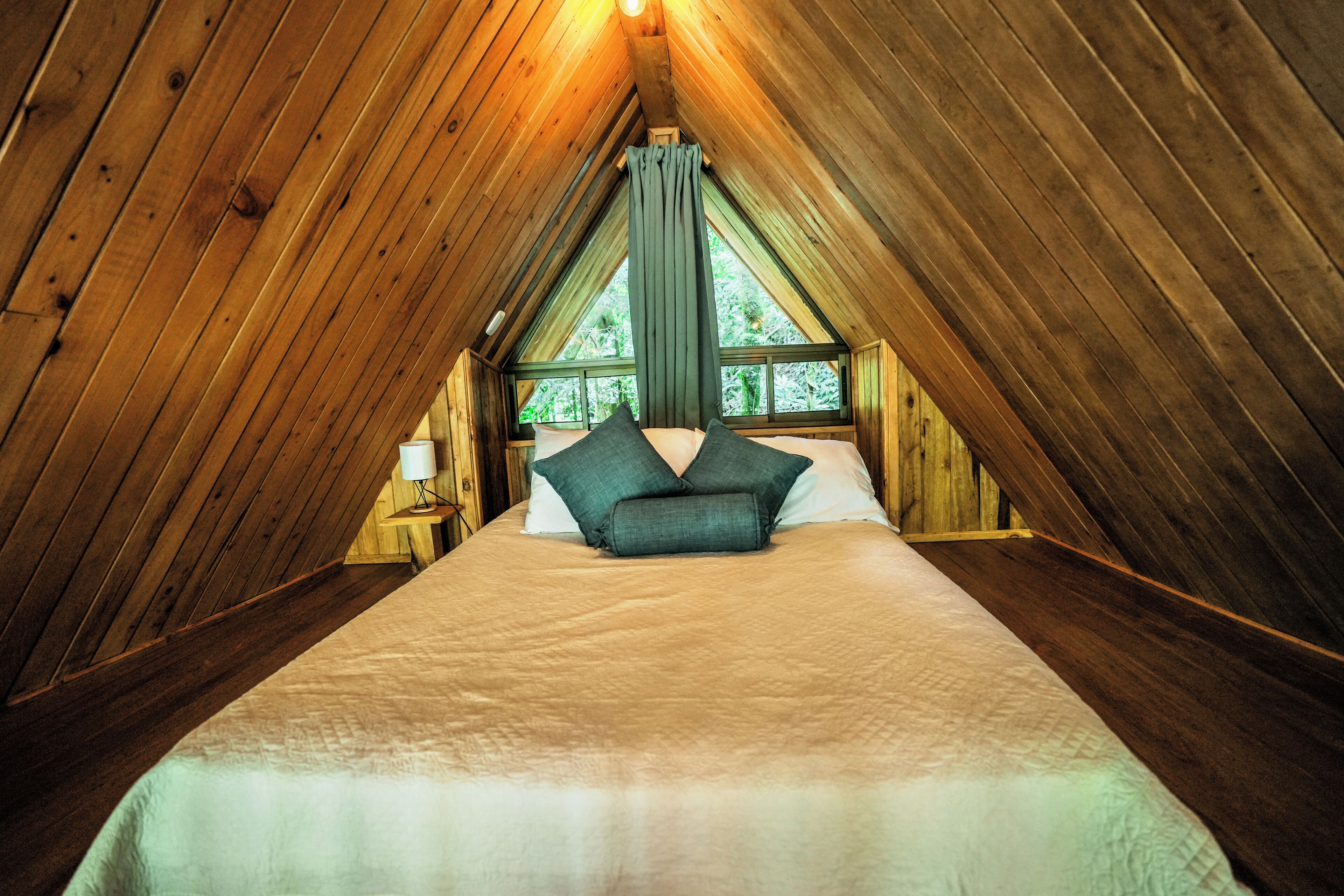 A bedroom with a wooden sloped ceiling, a bed with white bedding and green pillows, and a window with curtains.