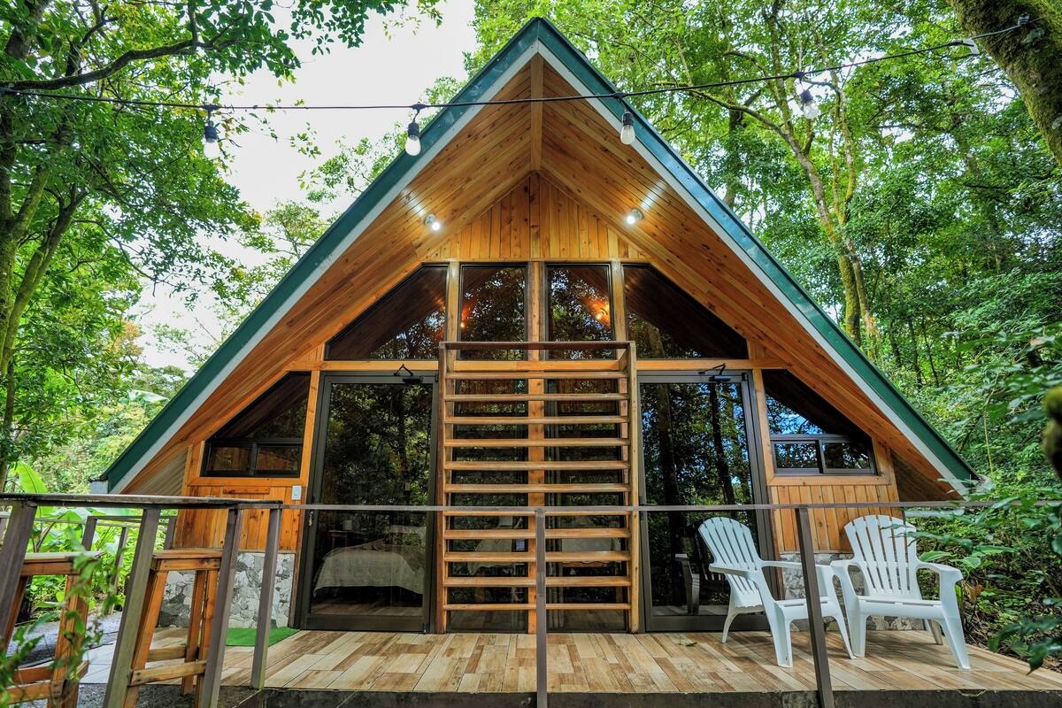 A wooden cabin with a sloped roof, large windows, and a wooden deck with chairs.
