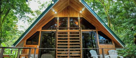 A wooden cabin with a sloped roof, large windows, and a wooden deck with chairs.
