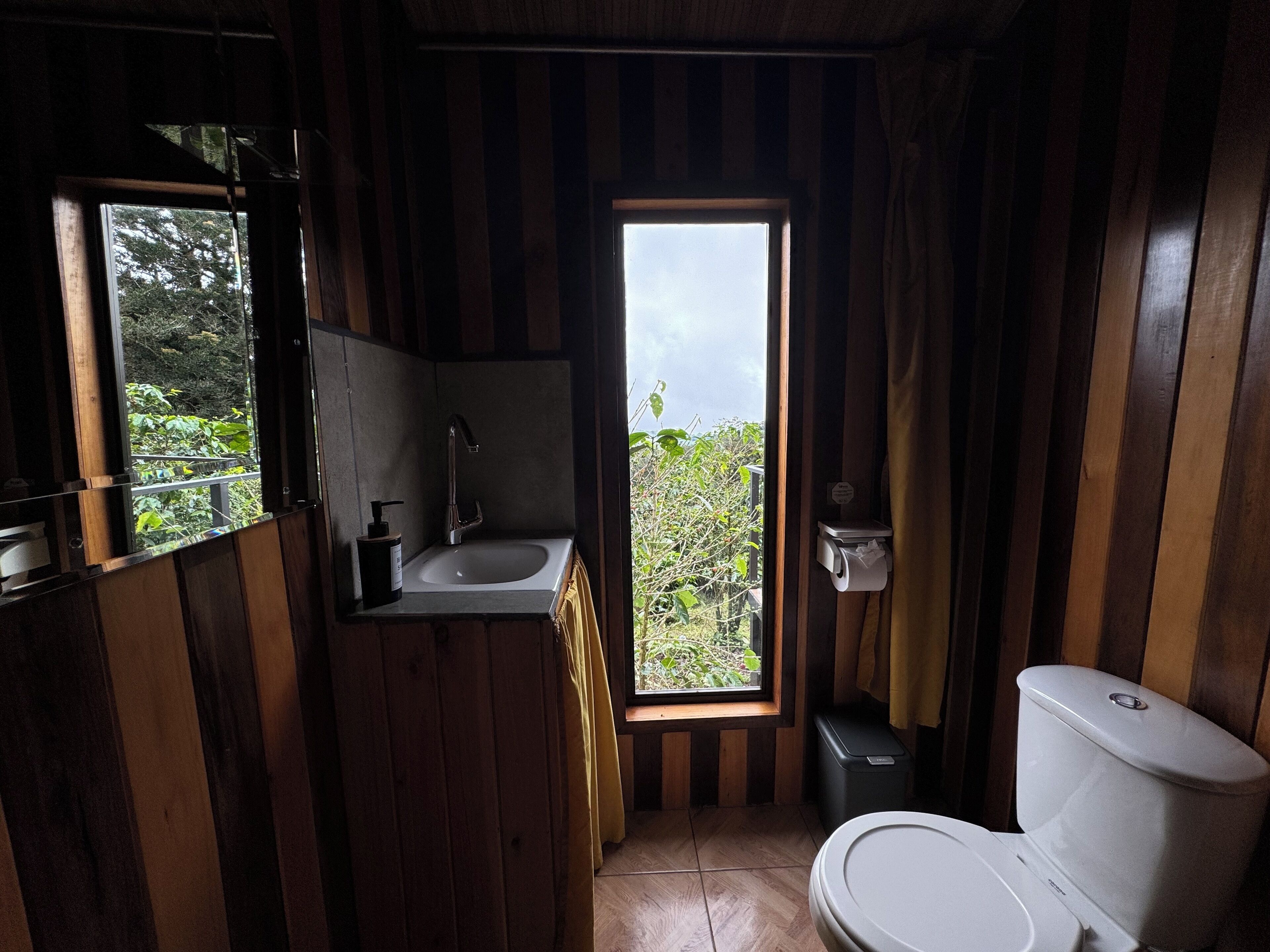 A compact bathroom with a sink, toilet, and a window providing a view of greenery outside.