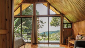 A spacious room with wooden floors, a large window, a bed, a sofa, and a small table. - Tityra Lodge (Monteverde)